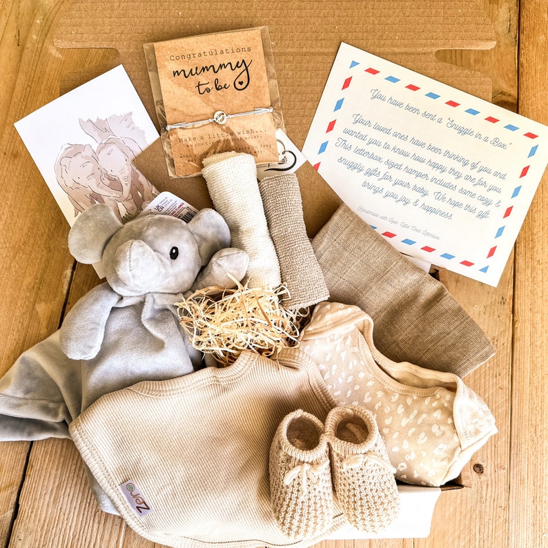 May include: A gift box filled with baby items, including a gray elephant plush toy, a beige knitted hat, a beige knitted onesie with a brown animal print, and a pair of beige knitted booties. The box also includes a card that reads "You have been sent a Snuggle in a Box."