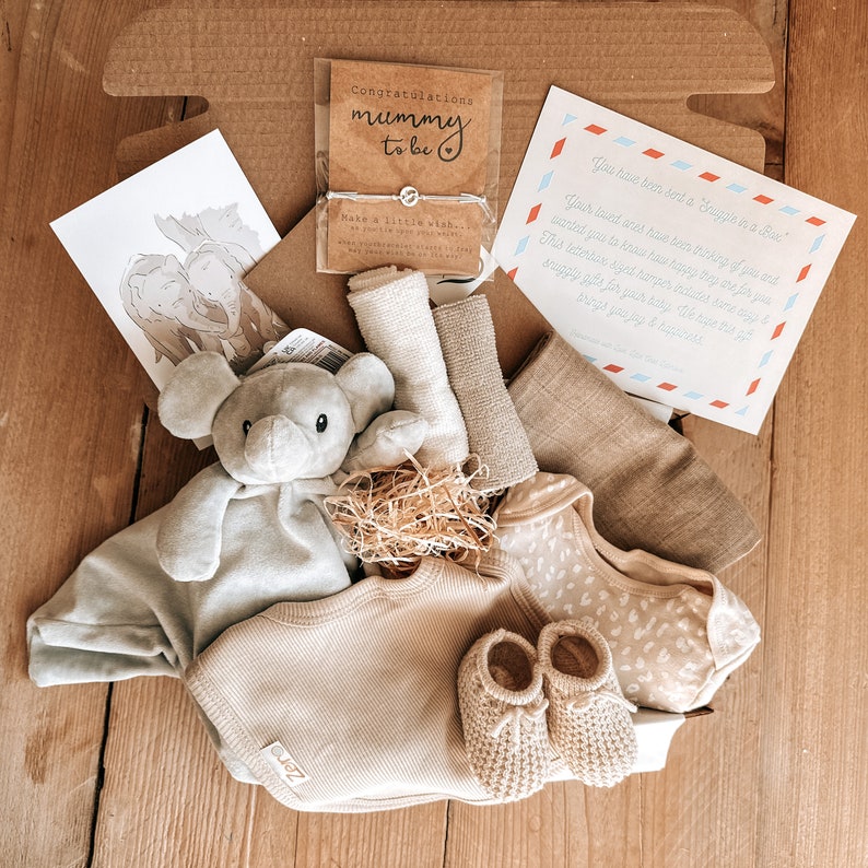 May include: A gift box filled with baby items, including a plush gray elephant toy, a beige knitted onesie, a pair of beige knitted booties, and a card that reads "Congratulations Mummy to be".