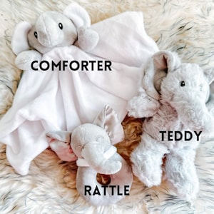 Puede incluir: Un conjunto de tres juguetes de peluche de elefante. El primero es una manta blanca con una cabeza de elefante gris unida. El segundo es un oso de peluche de elefante gris. El tercero es un sonajero de elefante gris.