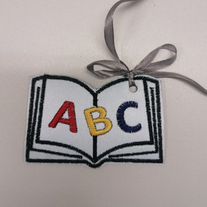 Puede incluir: Un adorno blanco en forma de libro con un contorno negro y las letras ABC bordadas en rojo, amarillo y azul. El adorno tiene un agujero perforado en la parte superior y una cinta gris atada alrededor.