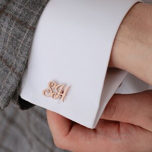 May include: Rose gold cufflinks with a monogram design. The cufflinks are worn on a white dress shirt with a gray plaid suit jacket.