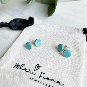 May include: A pair of teal blue stud earrings with silver posts. The earrings are in a small white pouch with the text "Mari Fiona Jewellery" printed on it.