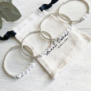 May include: Four silver beaded bracelets with white letter beads spelling out the names "Teddy", "Sam", "Stephen", and "Jo". The bracelets are displayed on a white fabric pouch with the text "Mari & Friends Jewellery".
