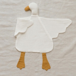 May include: A white goose-shaped baby toy made of soft, textured fabric. The toy features a yellow beak and feet, with outstretched wings. The toy is designed to be a comforting and engaging item for infants.