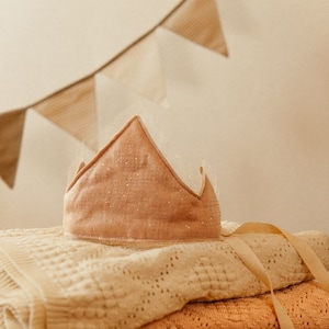 May include: A pink crown with glitter accents sits on top of a stack of beige and peach blankets. The blankets are folded and arranged on a brown fuzzy surface.