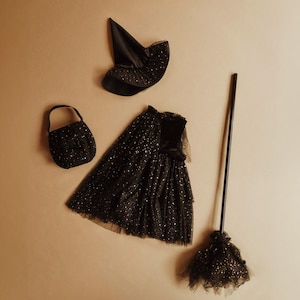 Può includere: Un set di costume da strega nero per bambini, che comprende un vestito, un cappello, una scopa e una piccola borsa. Il vestito è in tulle nero con glitter dorati e il cappello è in feltro nero con una punta a punta.