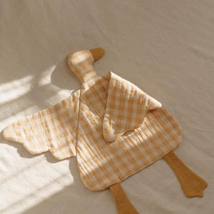 May include: A soft, yellow and white checkered duck-shaped baby comforter. The comforter has a head, wings, and feet, all in a matching yellow and white gingham pattern. The duck's beak and feet are a solid yellow color.