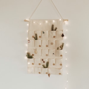 May include: A white canvas advent calendar with 24 pockets, decorated with small Christmas ornaments and greenery. The calendar is hanging on a white wall with a string of fairy lights.