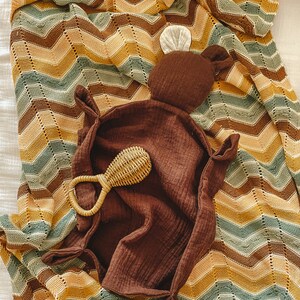 K&ouml;nnte beinhalten: Eine braune gestrickte Babydecke mit einem Zickzackmuster in Braun-, Gelb- und Gr&uuml;nt&ouml;nen. Ein braunes Pl&uuml;schtier mit einem wei&szlig;en Ohr liegt auf der Decke, zusammen mit einem geflochtenen Rassel.