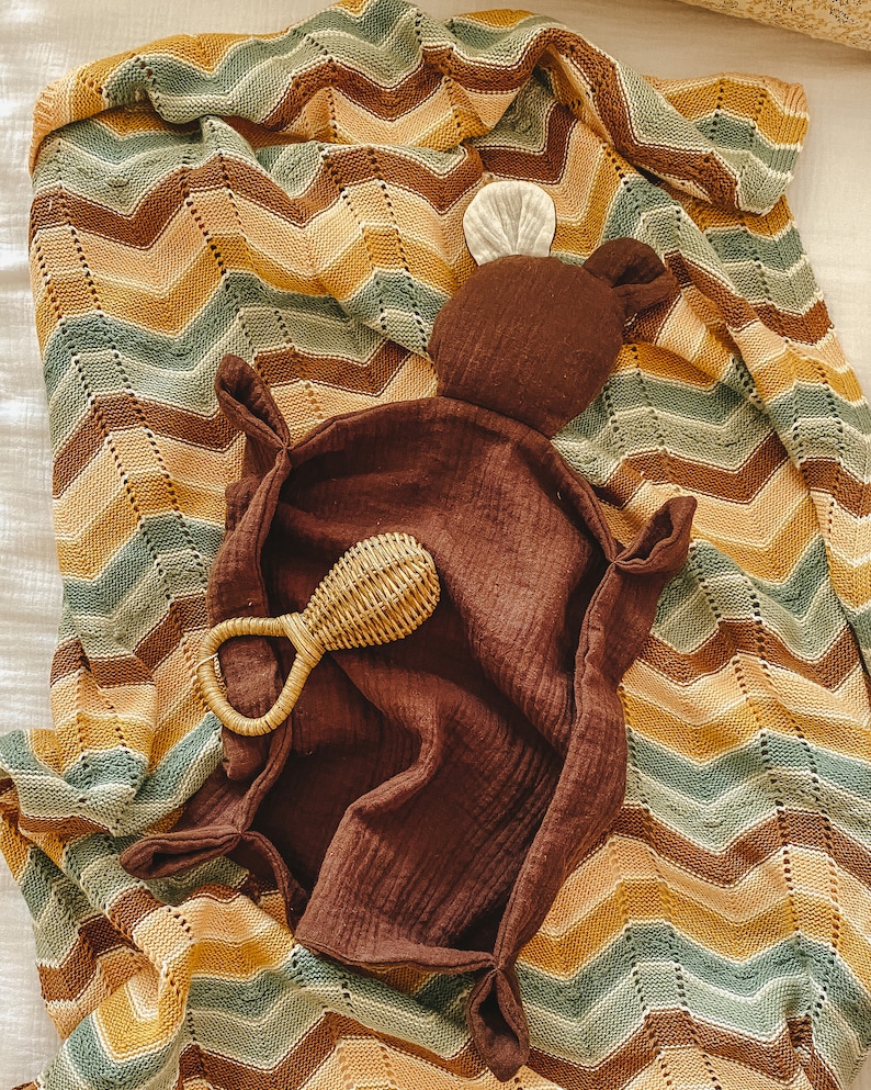 K&ouml;nnte beinhalten: Eine braune gestrickte Babydecke mit einem Zickzackmuster in Braun-, Gelb- und Gr&uuml;nt&ouml;nen. Ein braunes Pl&uuml;schtier mit einem wei&szlig;en Ohr liegt auf der Decke, zusammen mit einem geflochtenen Rassel.