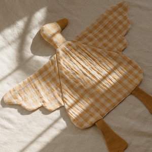 May include: A soft, yellow and white gingham patterned goose-shaped plush toy. The toy has outstretched wings and two yellow feet. The head and feet are a solid yellow color. The toy is lying on a white surface.