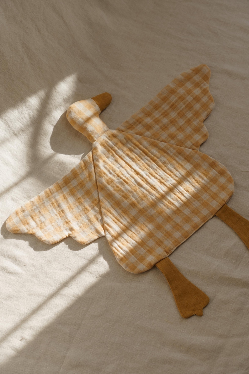 May include: A soft, yellow and white gingham patterned goose-shaped plush toy. The toy has outstretched wings and two yellow feet. The head and feet are a solid yellow color. The toy is lying on a white surface.