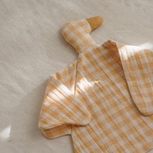 May include: A soft, yellow and white gingham duck-shaped baby blanket. The blanket has a textured appearance and is made of a lightweight fabric. The duck has a yellow beak and head, with the body forming the blanket.