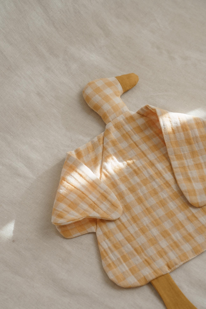 May include: A soft, yellow and white gingham duck-shaped baby blanket. The blanket has a textured appearance and is made of a lightweight fabric. The duck has a yellow beak and head, with the body forming the blanket.
