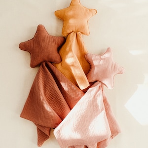 May include: Three star-shaped fabric pillows in shades of orange, pink, and brown are arranged in a triangular shape on a white background. The pillows are made of a soft, textured fabric and are perfect for a nursery or child's room.