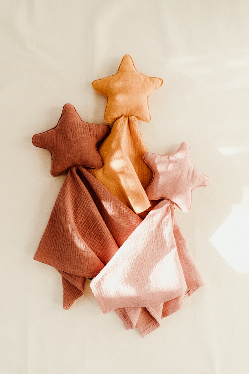 May include: Three star-shaped fabric pillows in shades of orange, pink, and brown are arranged in a triangular shape on a white background. The pillows are made of a soft, textured fabric and are perfect for a nursery or child's room.