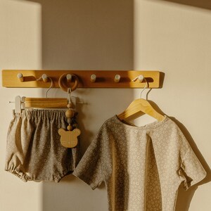 May include: A set of two baby clothes, a brown and white floral print top and matching bloomers, hanging on a wooden rack with a wooden bear rattle.