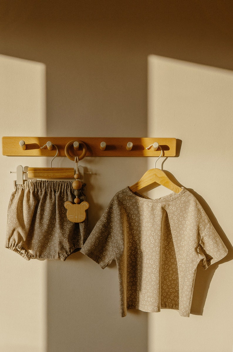 May include: A set of two baby clothes, a brown and white floral print top and matching bloomers, hanging on a wooden rack with a wooden bear rattle.