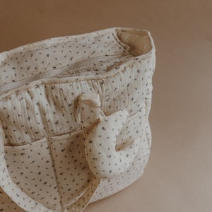 May include: A cream-colored diaper bag with a floral pattern and a small, cream-colored stuffed animal attached to the side.