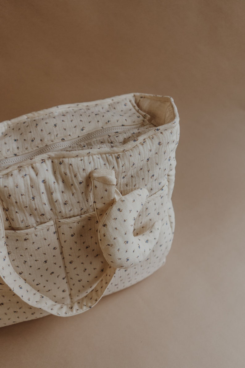 May include: A cream-colored diaper bag with a floral pattern and a small, cream-colored stuffed animal attached to the side.