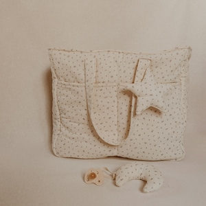 May include: A beige diaper bag with a floral pattern and a star-shaped stuffed toy. A pacifier and a crescent-shaped stuffed toy are in the foreground.