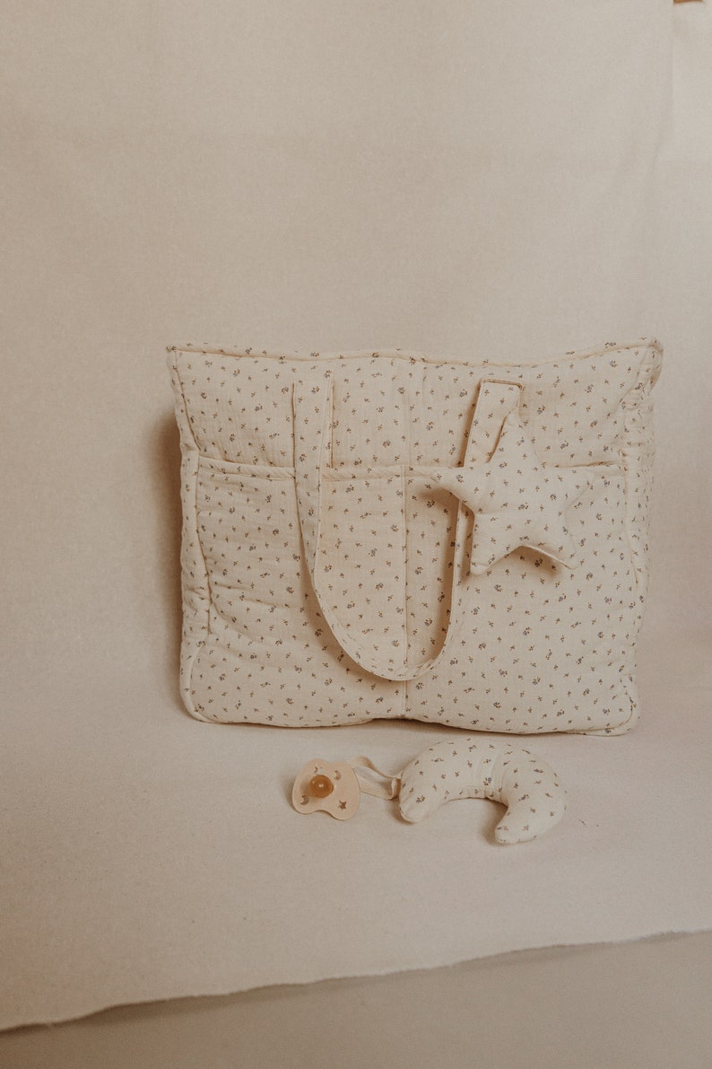 May include: A beige diaper bag with a floral pattern and a star-shaped stuffed toy. A pacifier and a crescent-shaped stuffed toy are in the foreground.