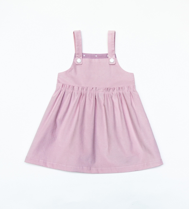 May include: A pink sleeveless dress with a gathered skirt and straps. The dress has white buttons on the straps and a white and purple patterned fabric on the bodice.