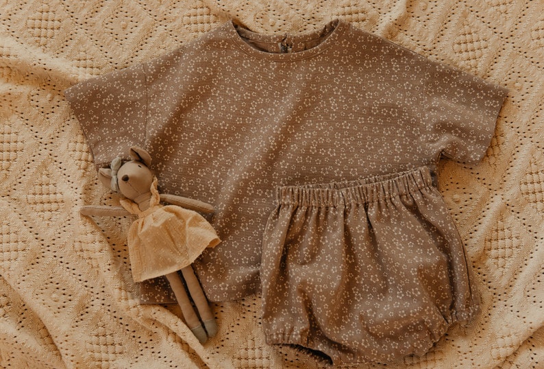 May include: A brown and white floral print baby outfit with a short-sleeved top and bloomers. A plush toy mouse is lying on the fabric.
