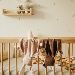 K&ouml;nnte beinhalten: Ein Holzbettchen mit einem wei&szlig;en Laken und einem braunen und rosafarbenen Stofftier, das an der Seite h&auml;ngt. Das Bettchen ist mit einer gelben und braunen Decke und einem Kissen dekoriert.