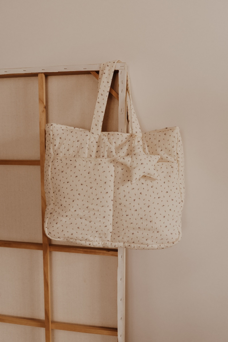 May include: A white tote bag with a small floral pattern and a star-shaped embellishment. The bag has a quilted texture and two handles.