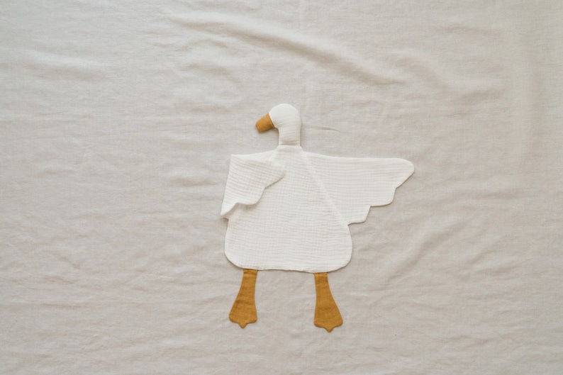 May include: A white goose-shaped baby toy made of soft, textured fabric. The toy features a yellow beak and feet, with outstretched wings. The toy is designed to be a comforting and engaging item for infants.