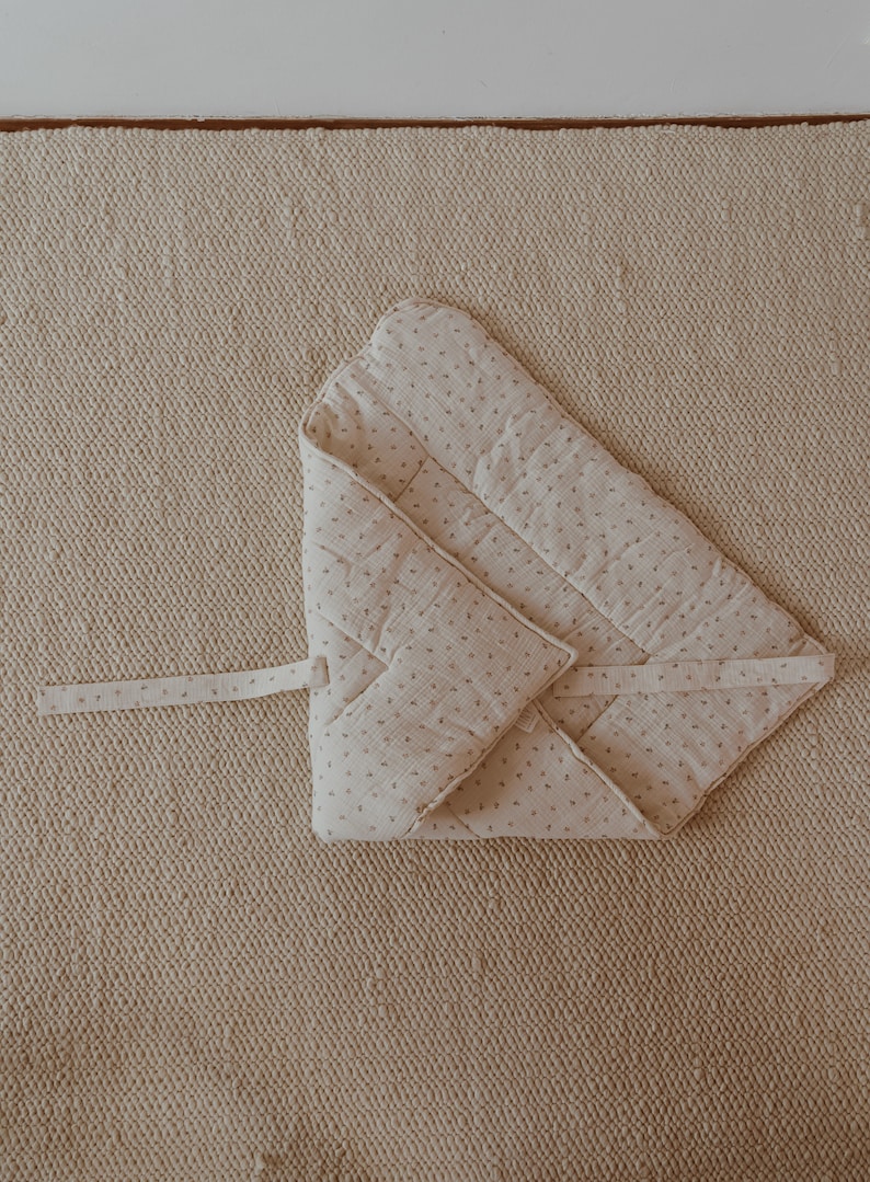 May include: A beige quilted baby blanket with a small floral pattern. The blanket is folded in half and tied with a ribbon.