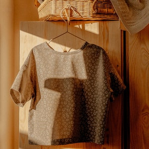 May include: A brown short-sleeved shirt with a white floral pattern hangs on a wooden hanger in front of a wooden closet door.