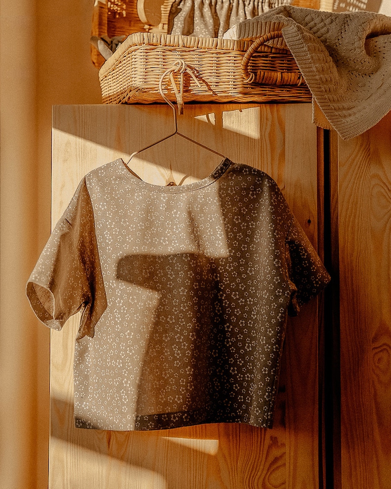 May include: A brown short-sleeved shirt with a white floral pattern hangs on a wooden hanger in front of a wooden closet door.