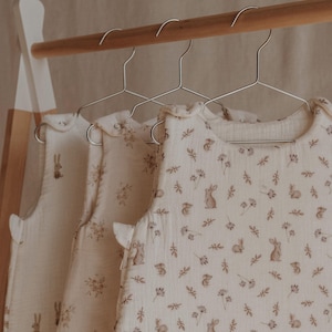 May include: Three white sleeveless baby sleep sacks with a brown bunny and leaf print hanging on a wooden rack.