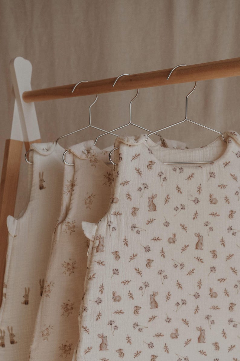 May include: Three white sleeveless baby sleep sacks with a brown bunny and leaf print hanging on a wooden rack.