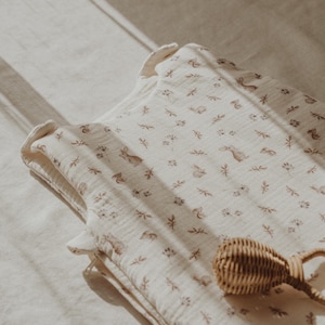 May include: A white muslin baby sleeping bag with a brown and white woodland animal print. The sleeping bag has a zipper closure and is lying on a white surface. A brown wicker rattle is also in the image.