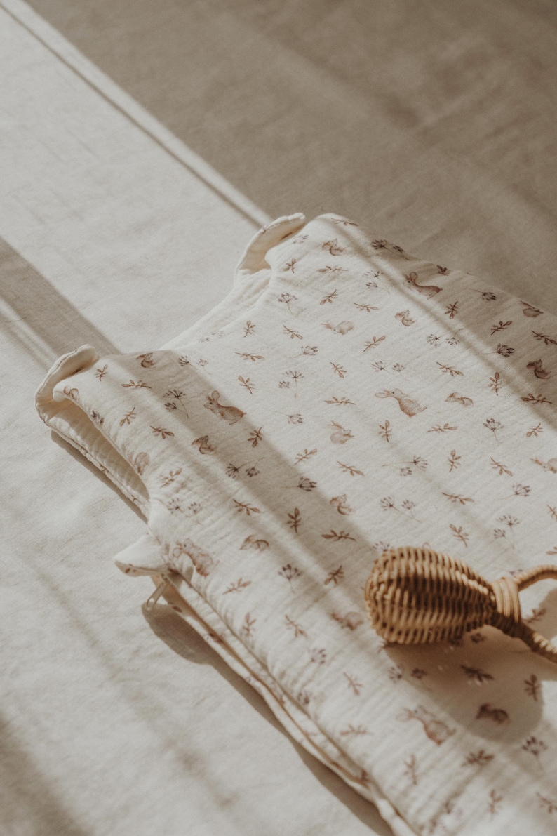 May include: A white muslin baby sleeping bag with a brown and white woodland animal print. The sleeping bag has a zipper closure and is lying on a white surface. A brown wicker rattle is also in the image.