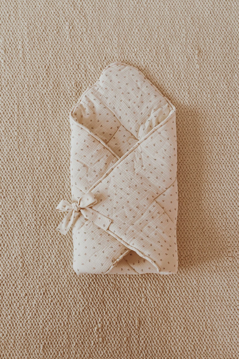 May include: A cream-colored swaddle blanket with a small floral pattern. The blanket is folded into a triangular shape and tied with a ribbon.