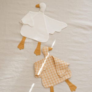 May include: Two soft, goose-shaped baby security blankets. One is white, the other is yellow gingham. Both have yellow beaks and feet. The blankets are made of a textured fabric and are laying on a neutral-colored surface.