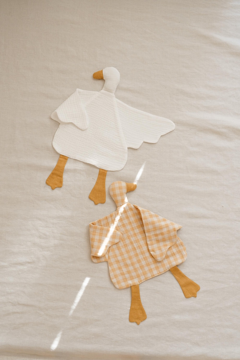 May include: Two soft, goose-shaped baby security blankets. One is white, the other is yellow gingham. Both have yellow beaks and feet. The blankets are made of a textured fabric and are laying on a neutral-colored surface.