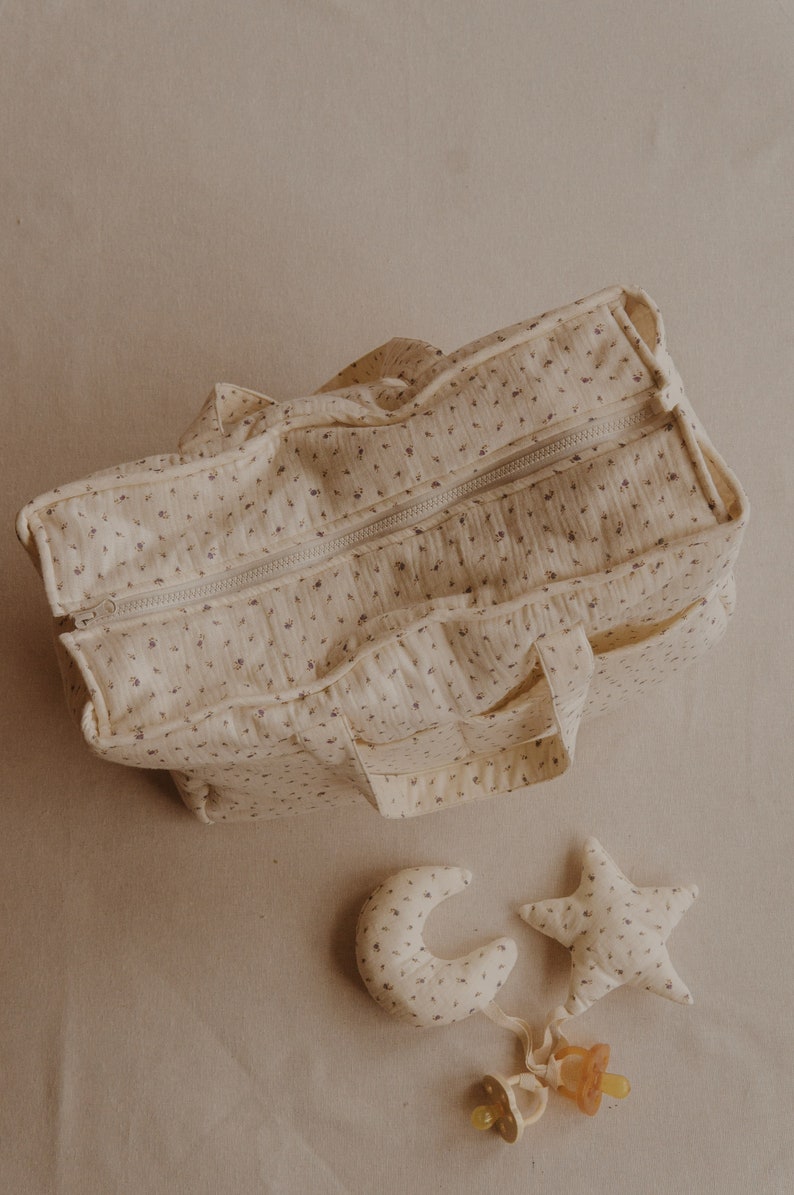 May include: A cream-colored diaper bag with a floral pattern and a zipper closure. The bag has two handles and multiple pockets. A matching cream-colored crescent moon and star with a pacifier attached are in the foreground.