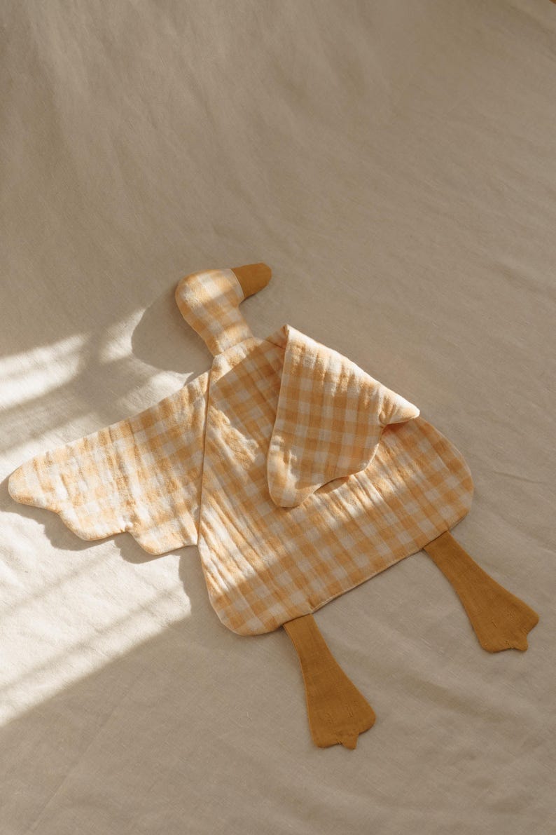 May include: A soft, yellow and white checkered duck-shaped baby comforter. The comforter has a head, wings, and feet, all in a matching yellow and white gingham pattern. The duck's beak and feet are a solid yellow color.
