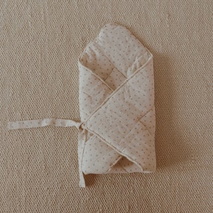May include: A white muslin swaddle with a small floral print, folded and tied with a ribbon. The swaddle is on a beige textured surface.