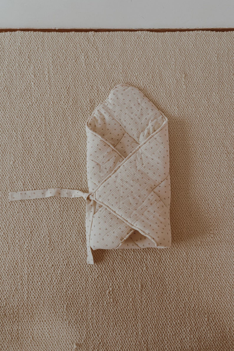May include: A white muslin swaddle with a small floral print, folded and tied with a ribbon. The swaddle is on a beige textured surface.