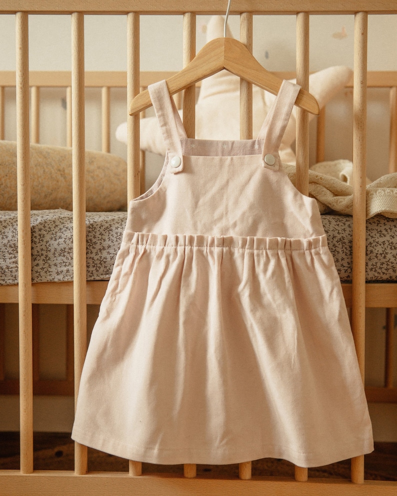 May include: A light pink linen overall dress with white buttons hanging on a wooden hanger in front of a wooden crib.