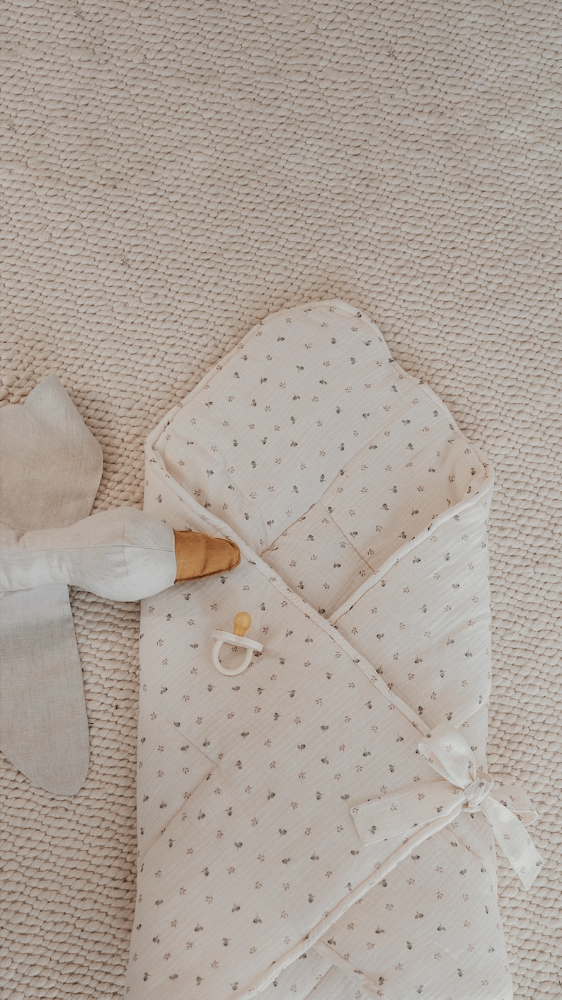 May include: A white swaddle blanket with a small floral pattern. The blanket is folded in a triangle shape and has a white ribbon tied around it. A wooden teether and a pacifier are lying on top of the blanket.