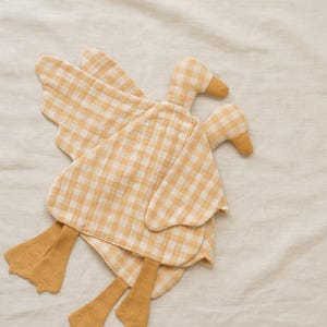 May include: Two yellow and white gingham duck-shaped stuffed toys with wings and feet. The ducks are made of soft fabric and are lying on a cream-colored surface. The toys have a simple, handmade aesthetic.