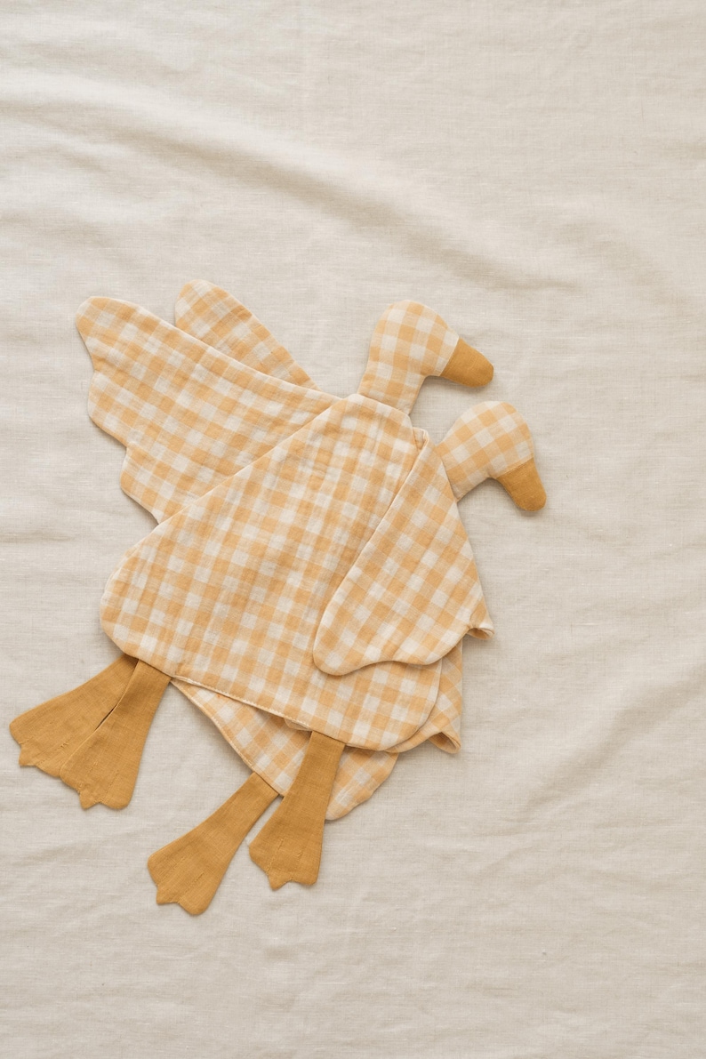 May include: Two yellow and white gingham duck-shaped stuffed toys with wings and feet. The ducks are made of soft fabric and are lying on a cream-colored surface. The toys have a simple, handmade aesthetic.