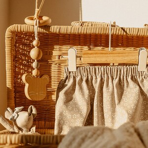 May include: A wicker basket with a wooden toy hanging from the top. The toy is a mouse with a ring and beads. Inside the basket is a pair of beige pants with a floral pattern and a beige knitted blanket.